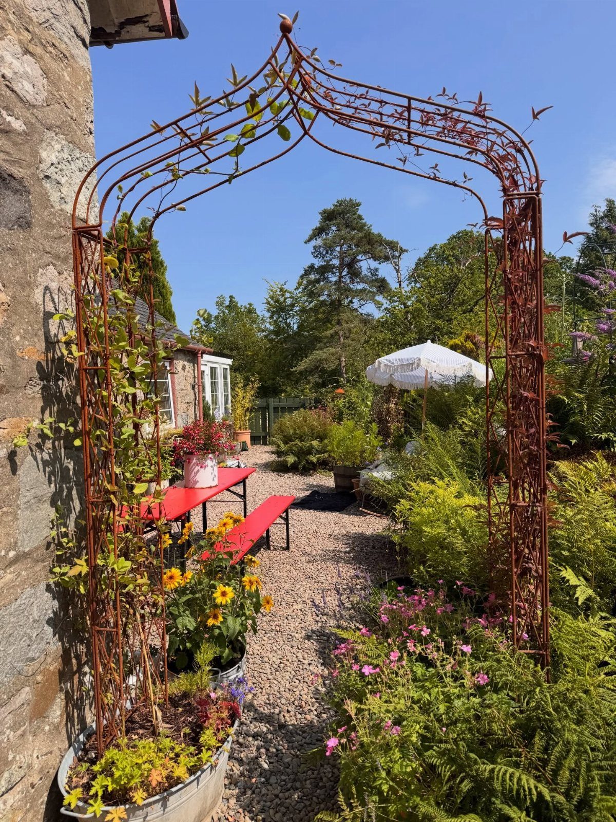 21 Back Garden Ideas from Potagers to Fire Pits 5 A rusty metal garden archway framing a gravel path and lush green garden plants.