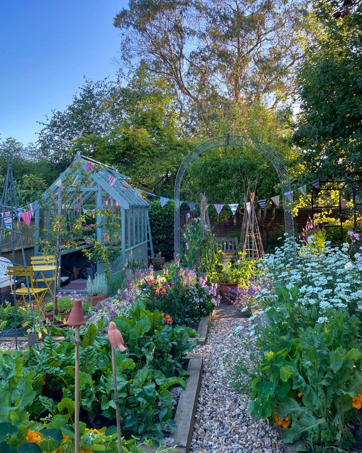 21 Back Garden Ideas from Potagers to Fire Pits 3 A vibrant garden with a greenhouse, gravel path, flowering plants, and decorative bunting under tall trees.
