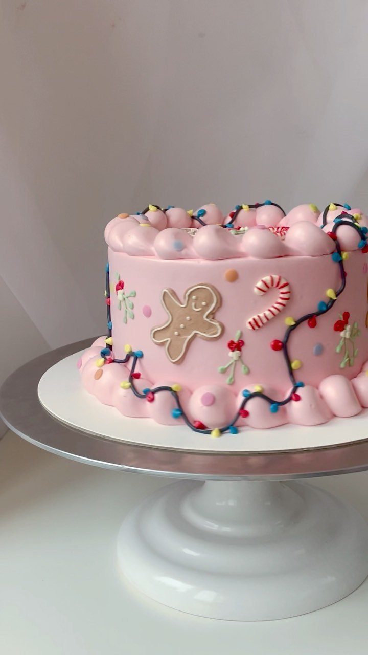 12 Beautiful Christmas Cake Ideas That Rival Bakery Work 11 Pink frosted cake decorated with gingerbread men, candy canes, and colorful icing lights on a stand.