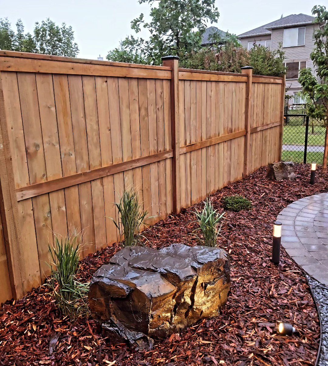 17 Cheap Yard Ideas for a Better Front or Back Garden 1 Dark reddish-brown bark mulch covers a garden bed along a wooden fence with plants and rocks.