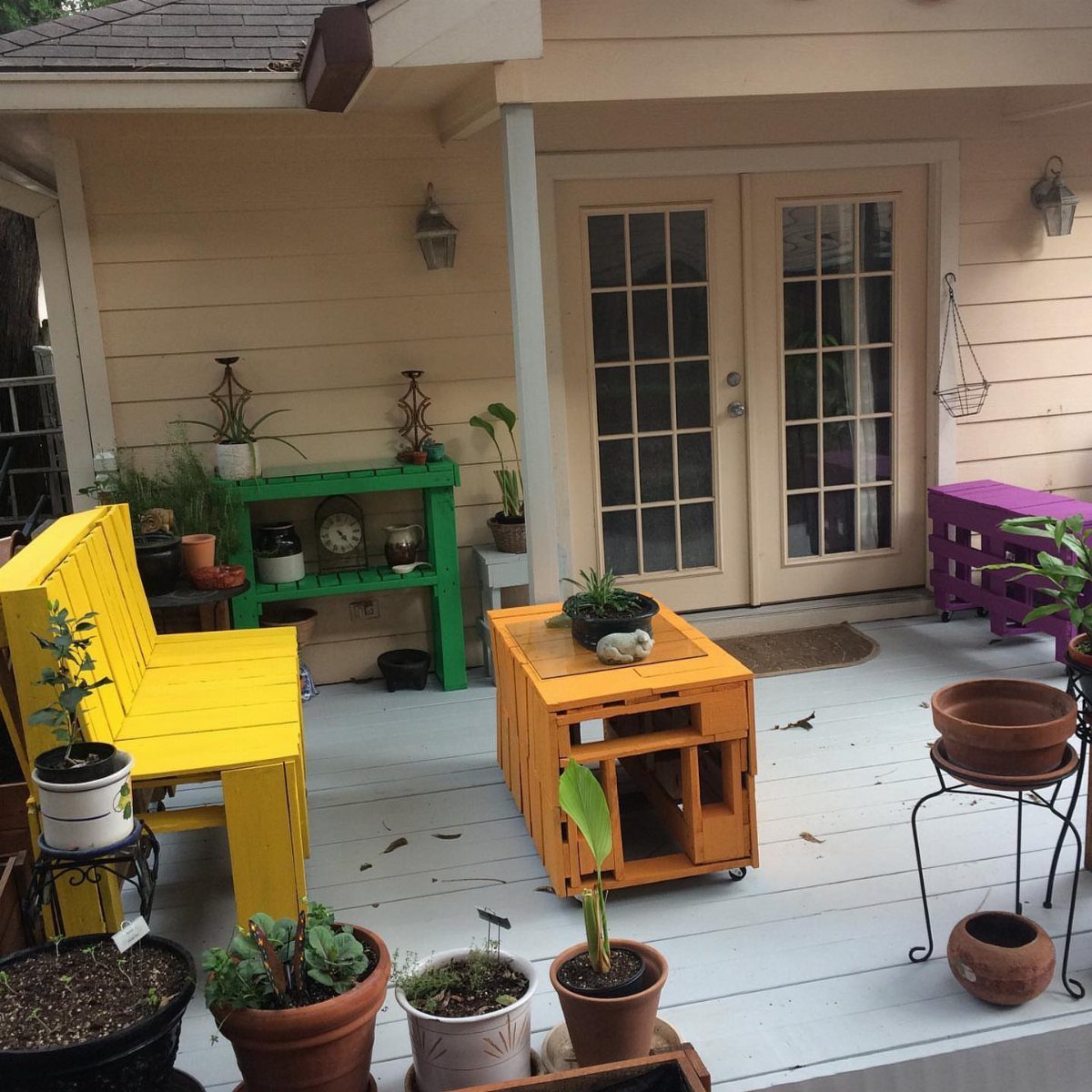 17 Cheap Yard Ideas for a Better Front or Back Garden 13 Brightly painted yellow, green, orange, and purple furniture pieces arranged on a white porch with potted plants.