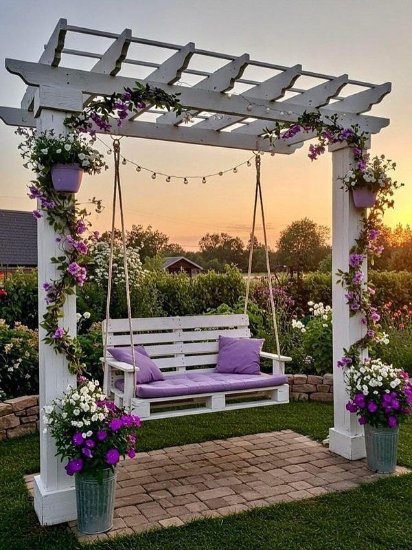 17 Cheap Yard Ideas for a Better Front or Back Garden 17 A white pallet swing with purple cushions hanging under a flower-decorated pergola at sunset.