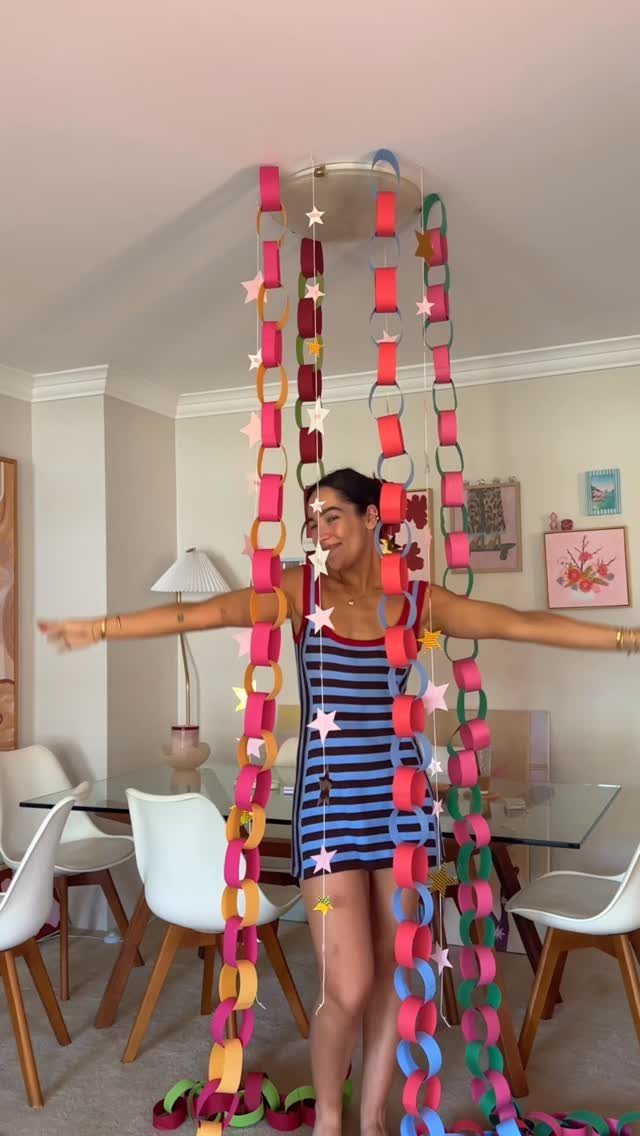 Christmas Decor Aesthetic: 13 Looks to Try This Season 9 A woman stands smiling amidst long, colorful hanging paper chains in a bright room.