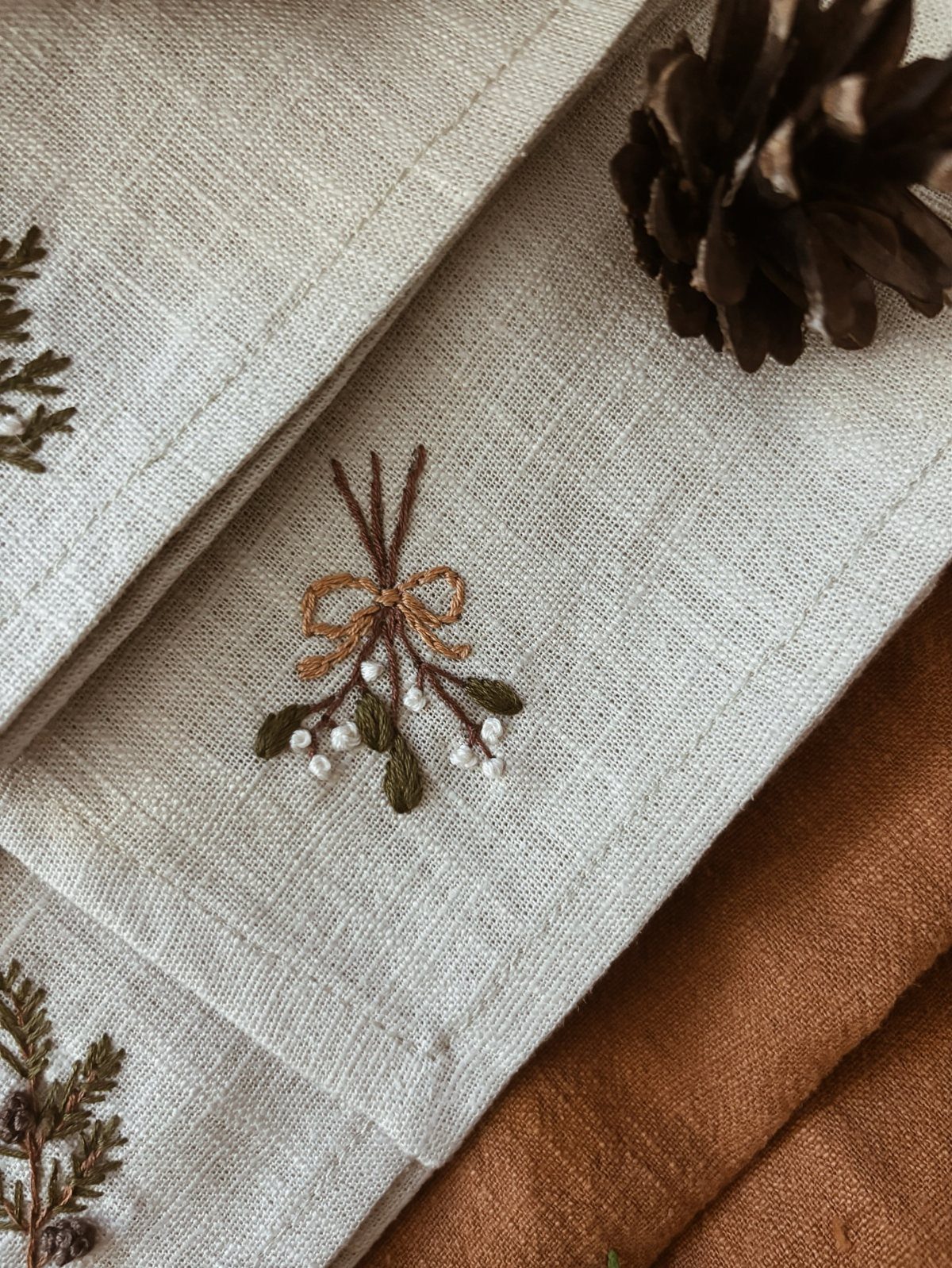Christmas Dinner and Table Decorations: 9 Ideas for Setting the Scene 9 Embroidered cream linen napkins with greenery sprigs and a dark pinecone on the side.