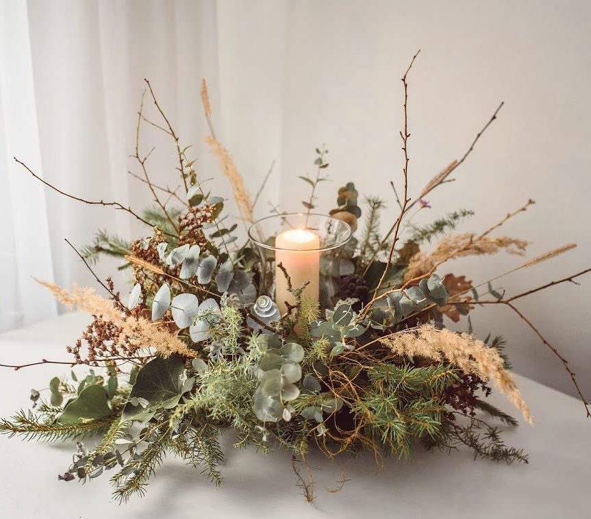 Christmas Dinner and Table Decorations: 9 Ideas for Setting the Scene 2 A festive centerpiece featuring greenery, eucalyptus, dried grasses, and a lit candle on a white table.
