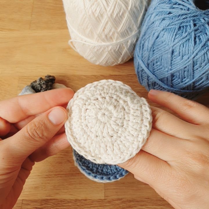 18 Christmas Gift Crochet Ideas for Everyone on Your List 9 Hands holding a white crocheted facial pad over a blue one on a wooden table.