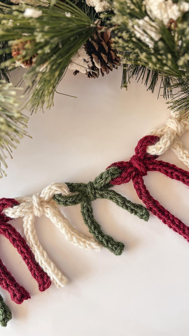 Knitted yarn garland with red, white, and green bows resting under pine branches.
