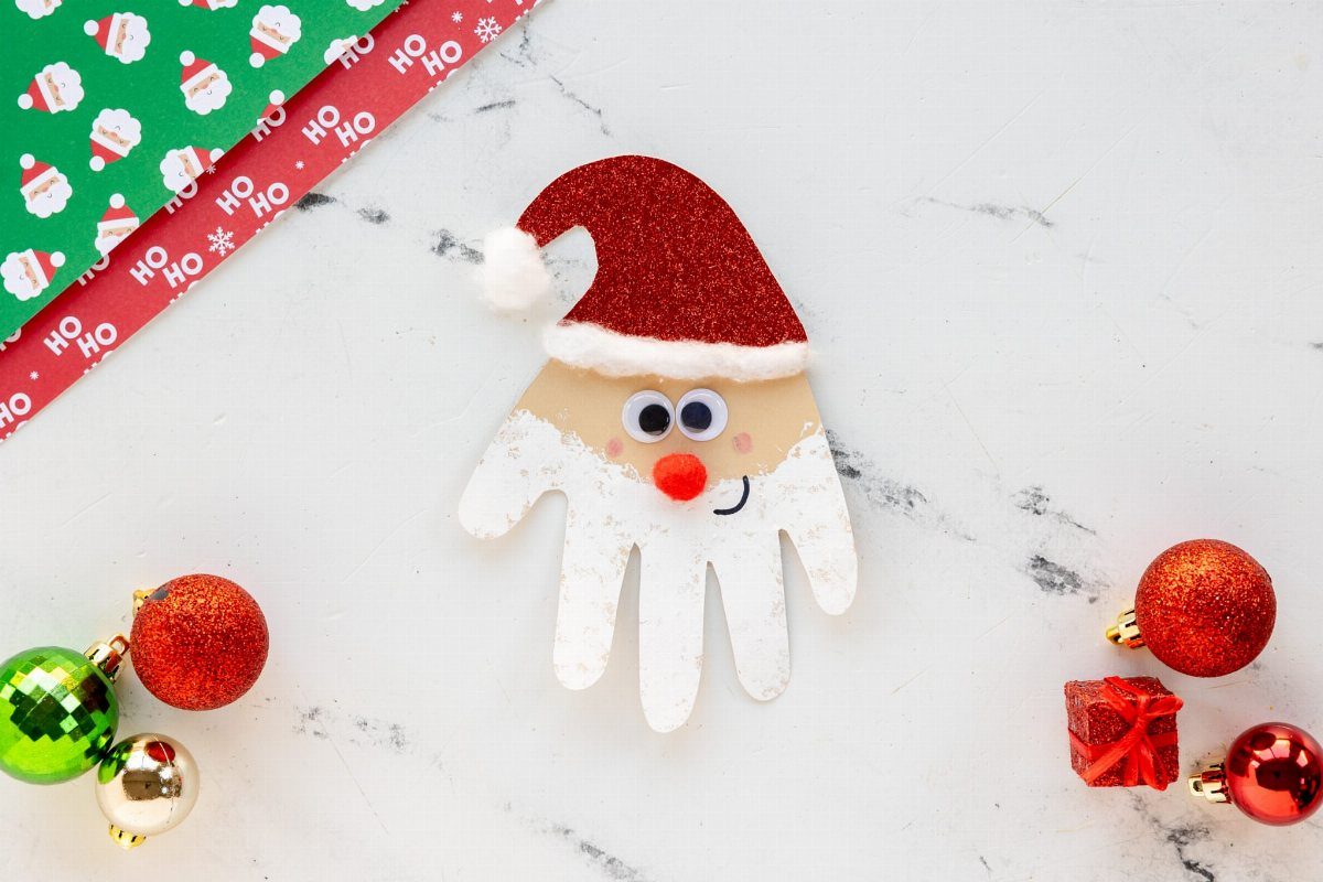 A white handprint cutout decorated as Santa Claus with a red glittery hat on marble.