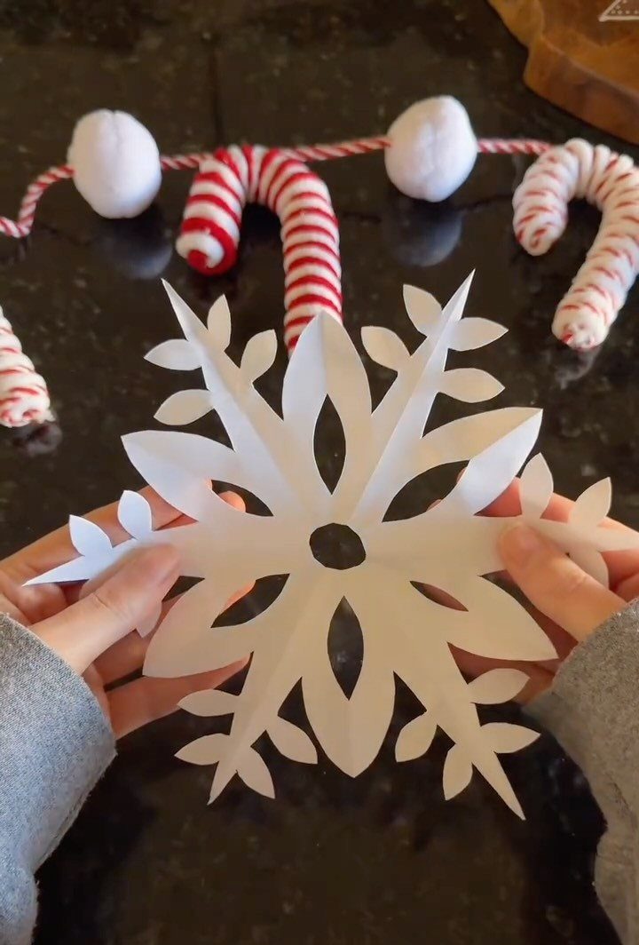 Hands holding a large, white, intricately cut paper snowflake decoration on a dark surface.