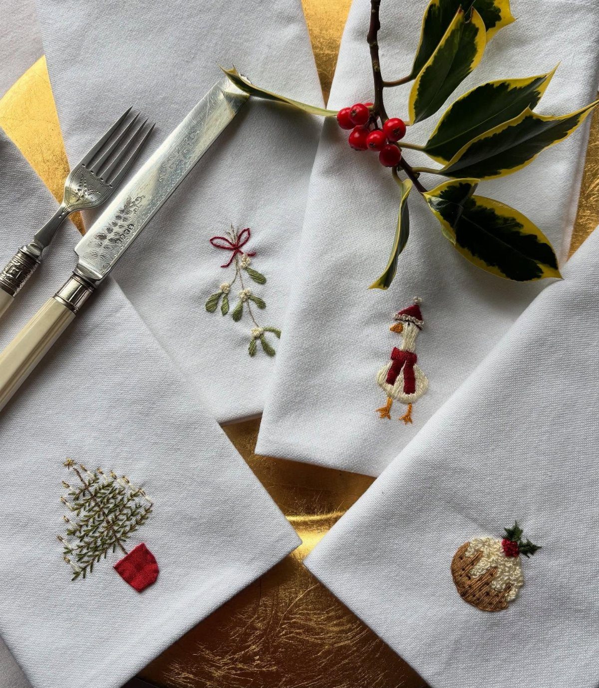 16 Christmas Sewing Projects That Cover the Whole Holiday 10 Embroidered white fabric napkins with festive holiday motifs arranged with silver cutlery and holly berries.