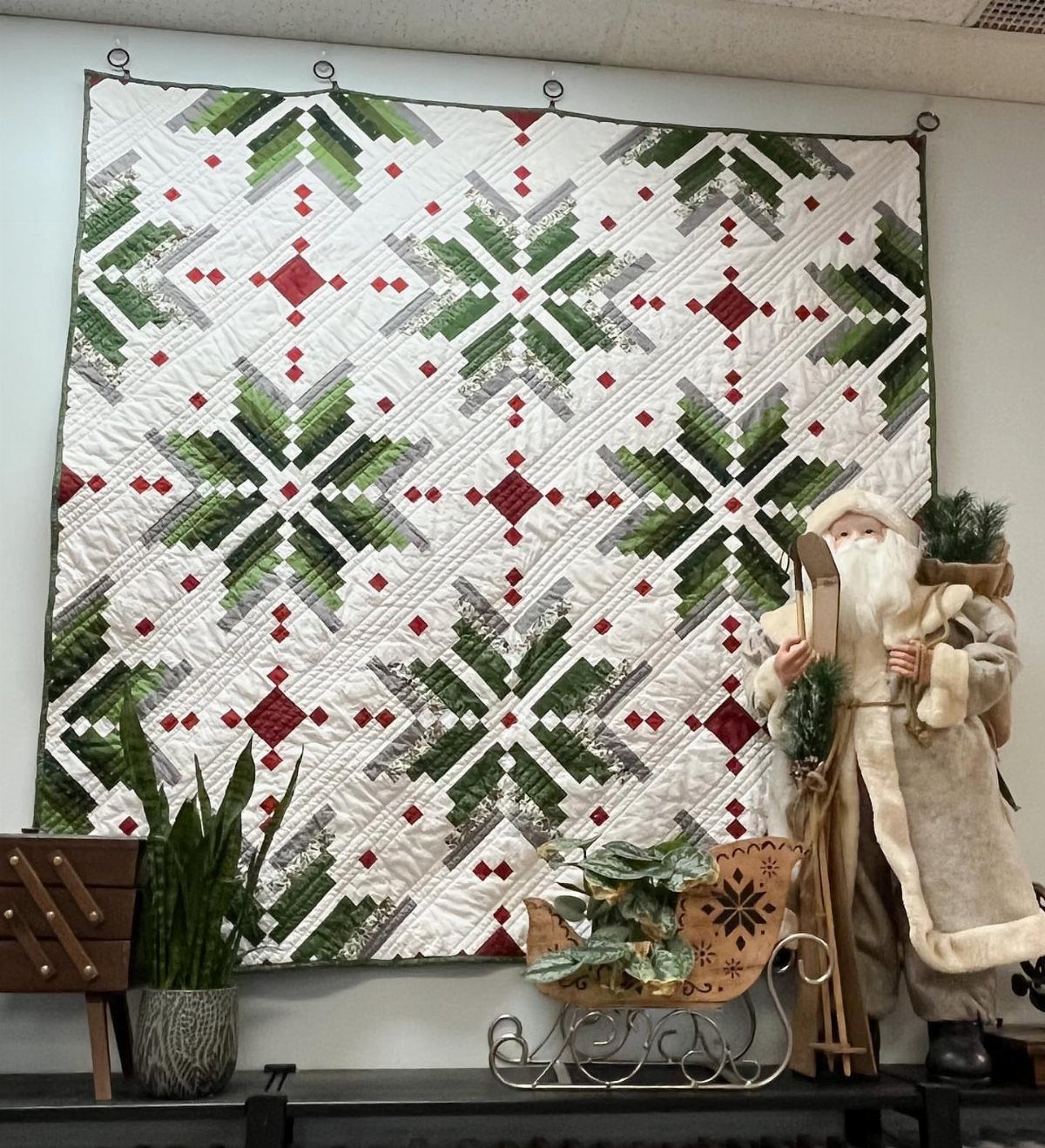 16 Christmas Sewing Projects That Cover the Whole Holiday 2 A white quilt with green and red star patterns hanging on a wall above festive decorations.