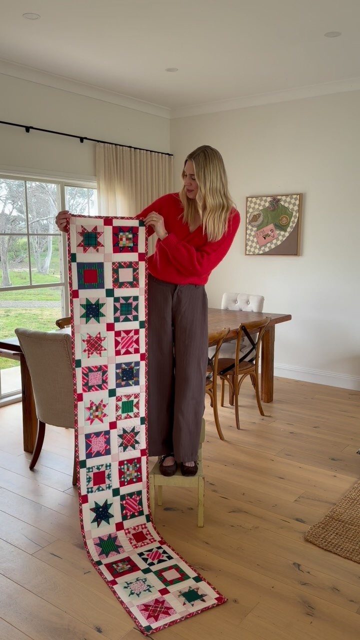 16 Christmas Sewing Projects That Cover the Whole Holiday 8 A woman holding up a long, colorful patchwork Christmas table runner in a dining room.