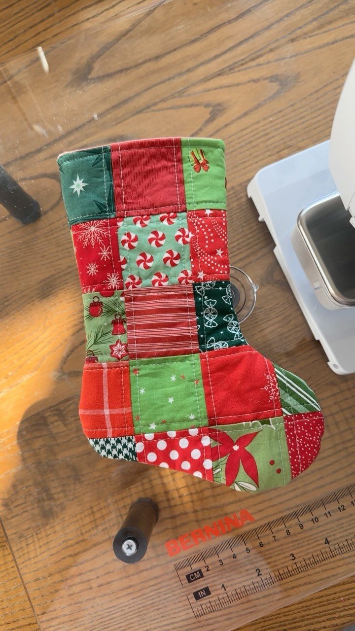 16 Christmas Sewing Projects That Cover the Whole Holiday 1 A small patchwork Christmas stocking resting on a wooden table near a sewing machine.