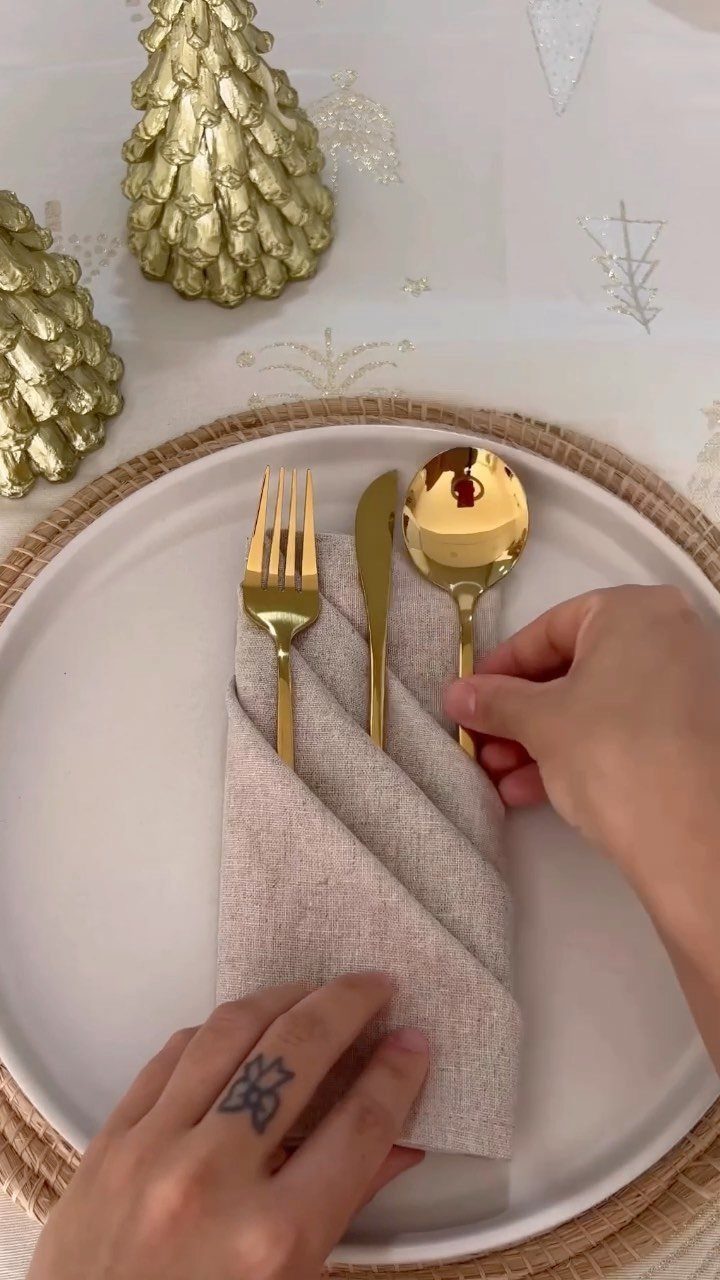 13 Christmas Table Decoration Ideas for Every Style 11 Hands holding a beige linen napkin with gold cutlery tucked into its folds on a white plate.