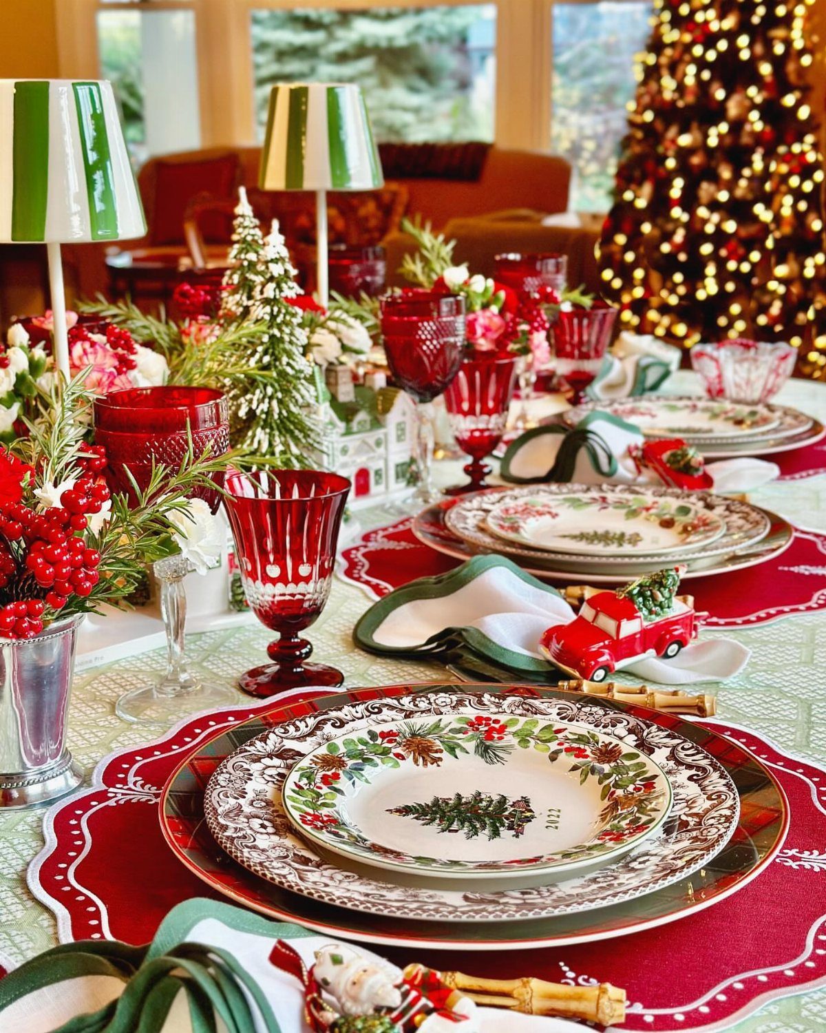 13 Christmas Table Decoration Ideas for Every Style 7 Christmas dinner plates with festive holly patterns set on a red tablecloth with red glassware.