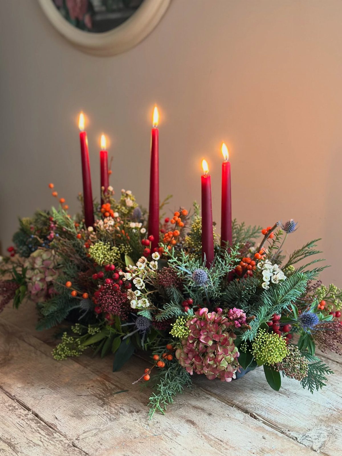 13 Christmas Table Decoration Ideas for Every Style 4 A festive floral centerpiece with lit red taper candles and various greenery on a wooden table.