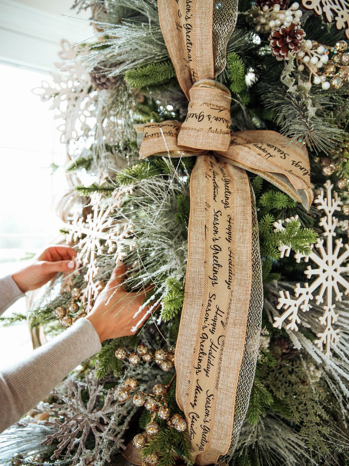 Christmas Tree Ribbon: 18 Styling Ideas That Work 7 Hands decorating a Christmas tree featuring a large burlap ribbon with holiday greetings and snowflake ornaments.