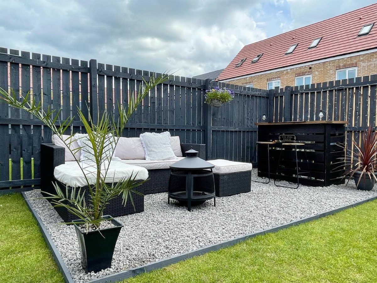 20 Corner Yard Garden Ideas for Any Size Space 12 Black wicker outdoor sofa and side table set on a grey gravel area surrounded by a dark fence.