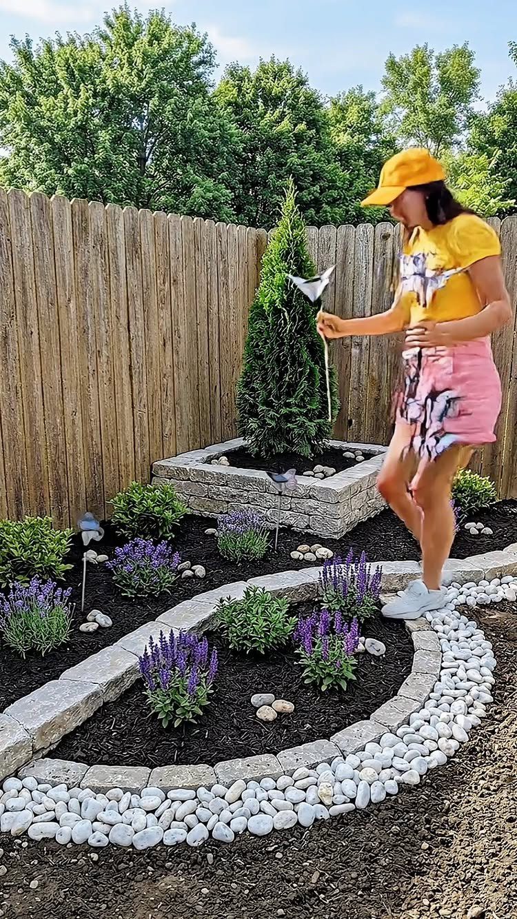 20 Corner Yard Garden Ideas for Any Size Space 1 A woman stands beside a stone raised garden bed with an evergreen tree in a backyard.
