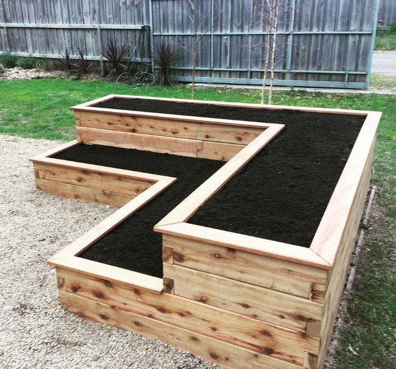 20 Corner Yard Garden Ideas for Any Size Space 3 A large L-shaped wooden tiered garden bed filled with dark soil on a gravel surface.