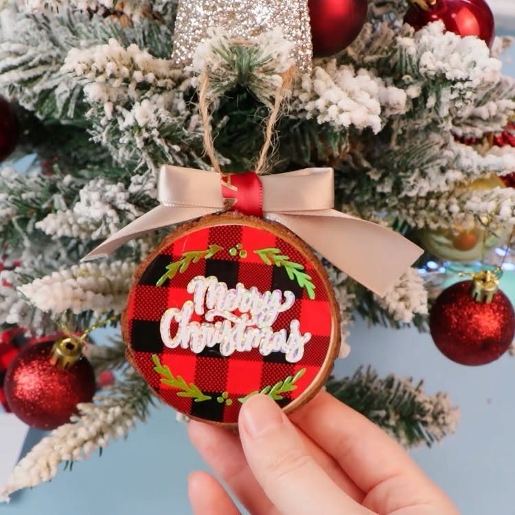 Cricut Christmas Balls: 14 DIY Ornaments for 2026 8 A hand holding a red and black buffalo check wooden Christmas ornament with white lettering.