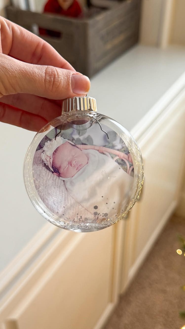 Cricut Christmas Balls: 14 DIY Ornaments for 2026 11 A hand holding a clear glass ornament featuring a printed photo of a sleeping newborn baby.