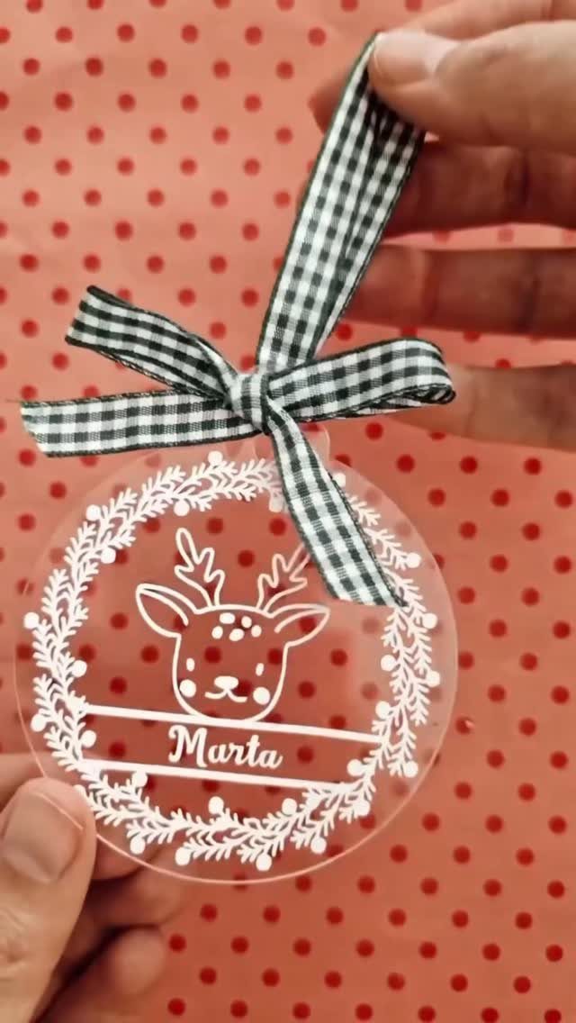 Cricut Christmas Balls: 14 DIY Ornaments for 2026 5 A clear acrylic ornament with a white vinyl reindeer and name held by hands.