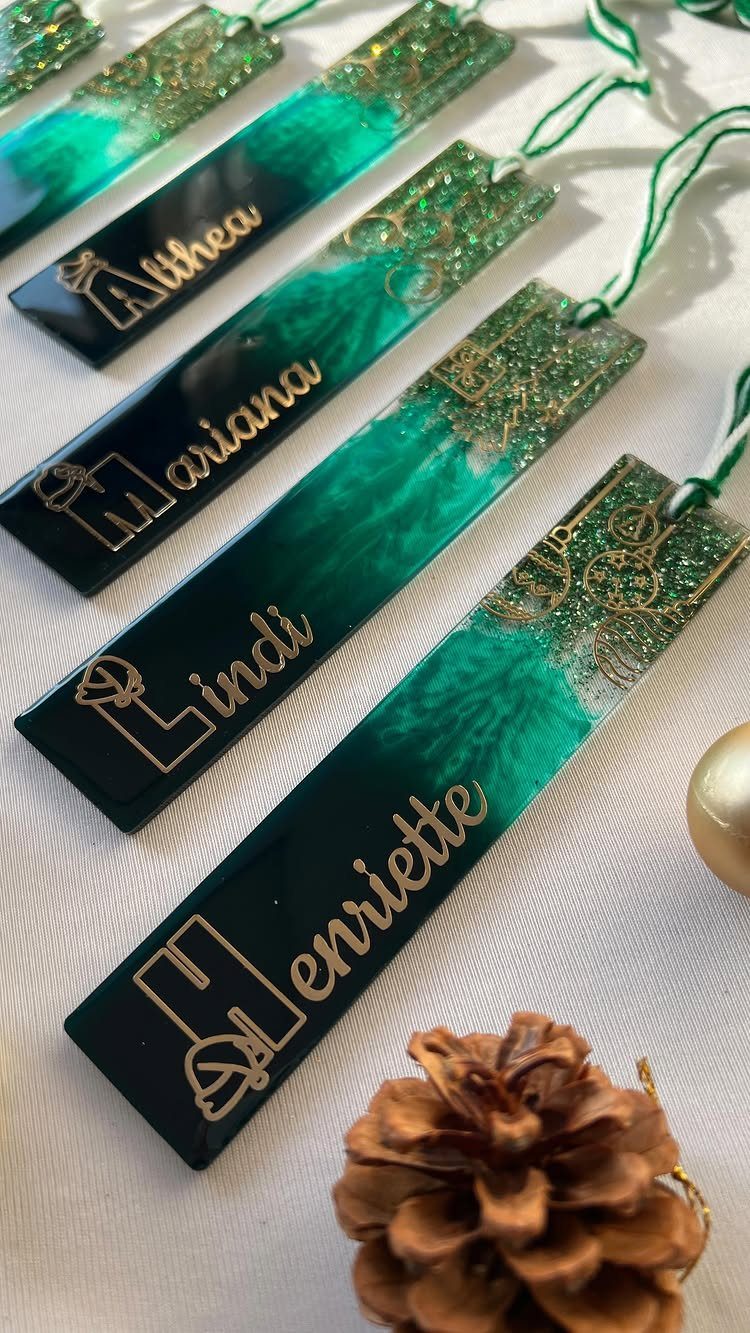 27 DIY Cheap Christmas Gifts for 2026 10 Resin bookmarks with green glitter and gold names laid out on a white surface.