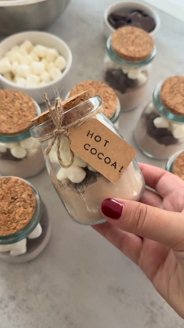 27 DIY Cheap Christmas Gifts for 2026 11 A hand holding a glass jar filled with layered hot cocoa mix and marshmallows.