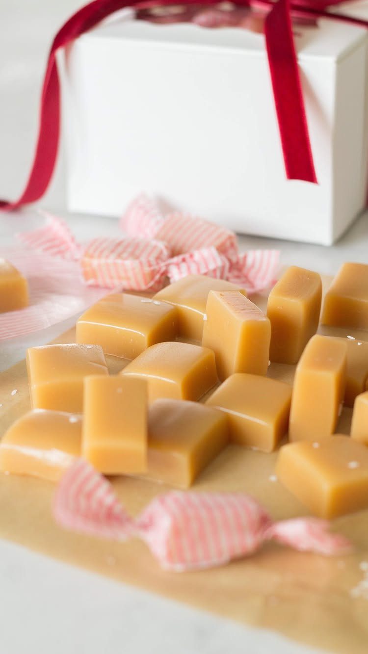 27 DIY Cheap Christmas Gifts for 2026 16 Golden brown caramel candies scattered on parchment paper near a white gift box.