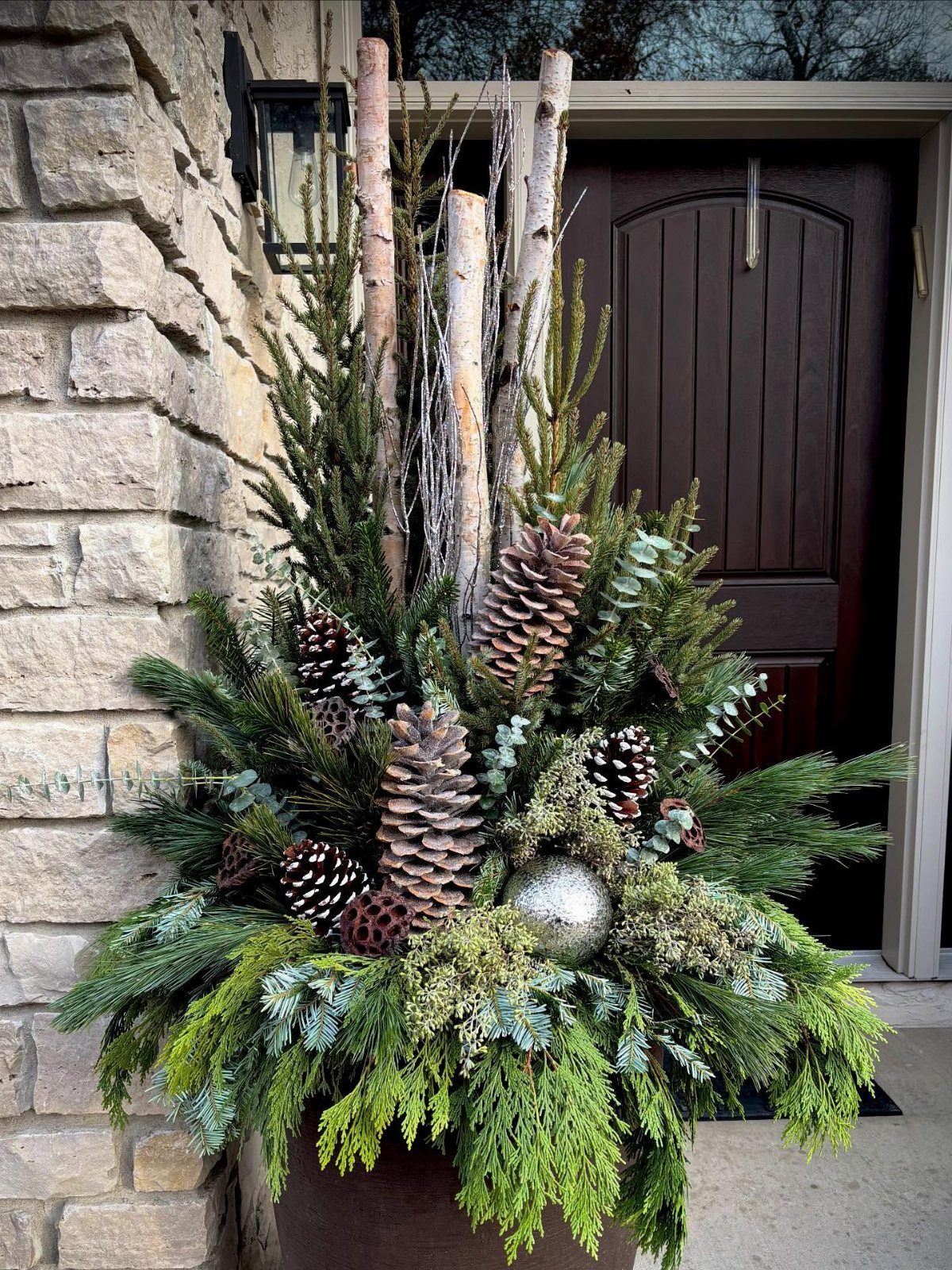A large planter filled with birch logs, cedar branches, eucalyptus, and pinecones near a stone wall.