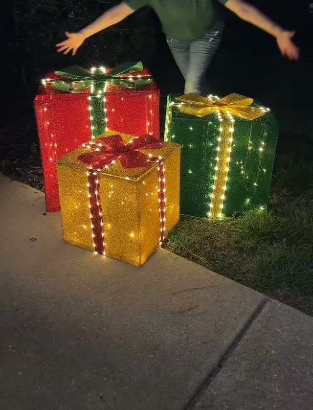 Three large, glowing, illuminated Christmas gift boxes with bows sitting on a dark lawn.