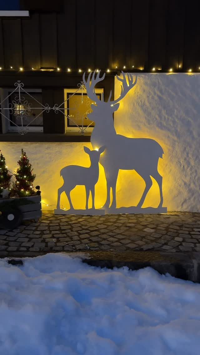 Two backlit white wooden reindeer silhouettes standing on a cobblestone path near snow.