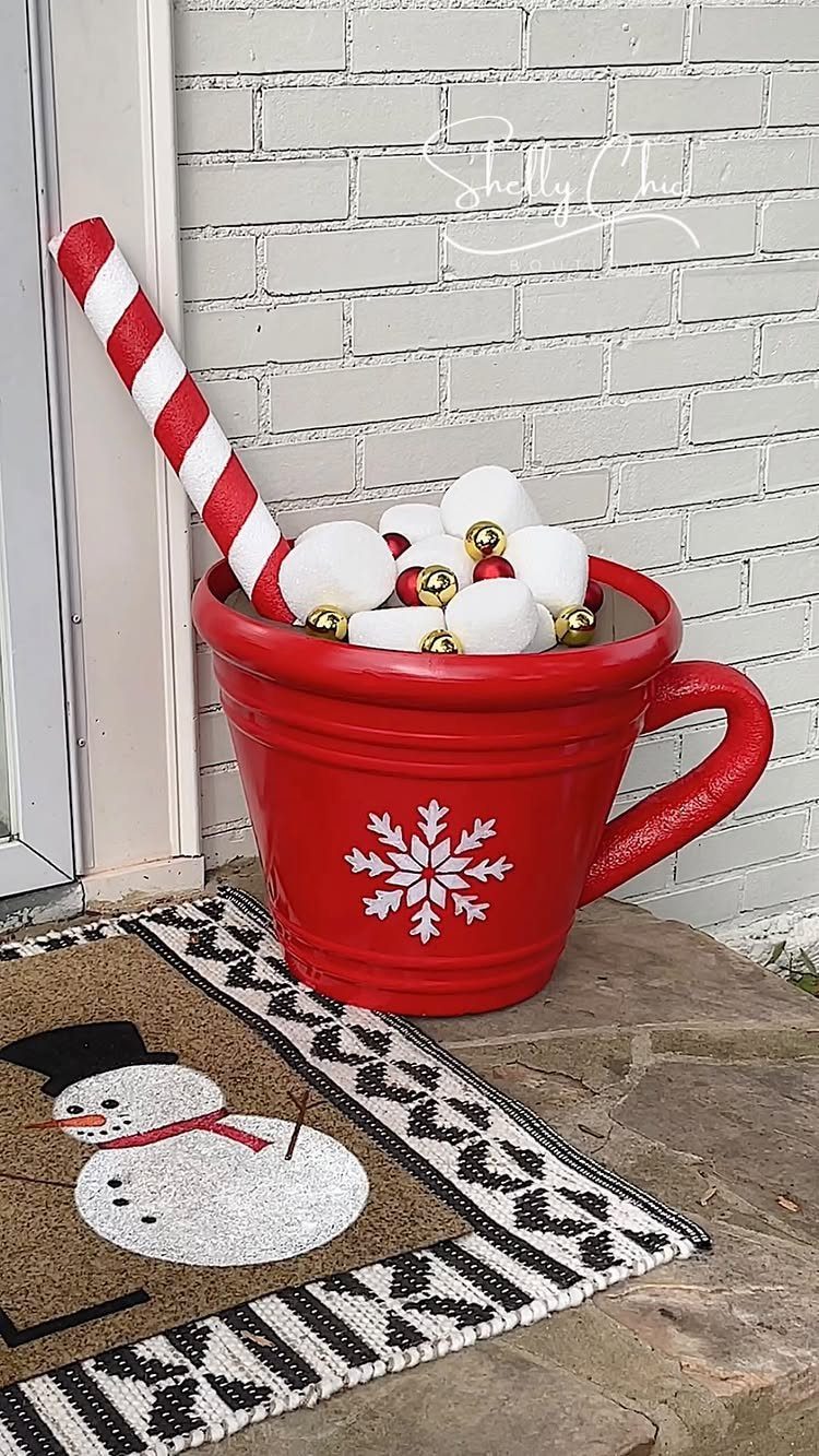 A large red mug planter with a snowflake design sitting on a porch next to a snowman mat.