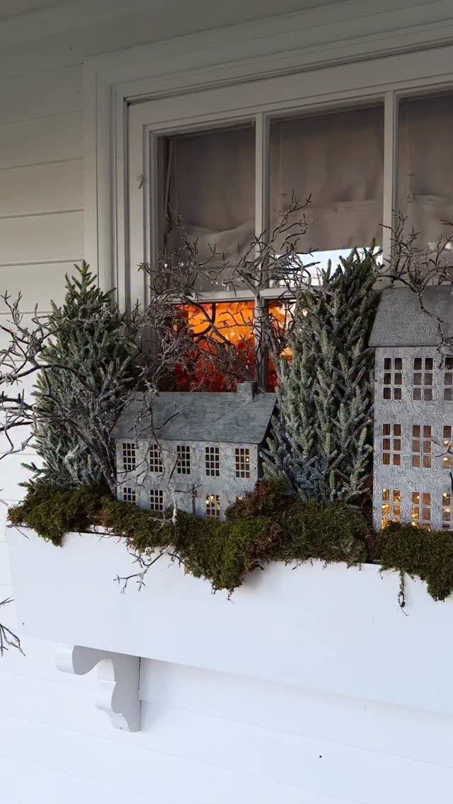 Small illuminated decorative houses and evergreen trees nestled in a mossy window box planter.