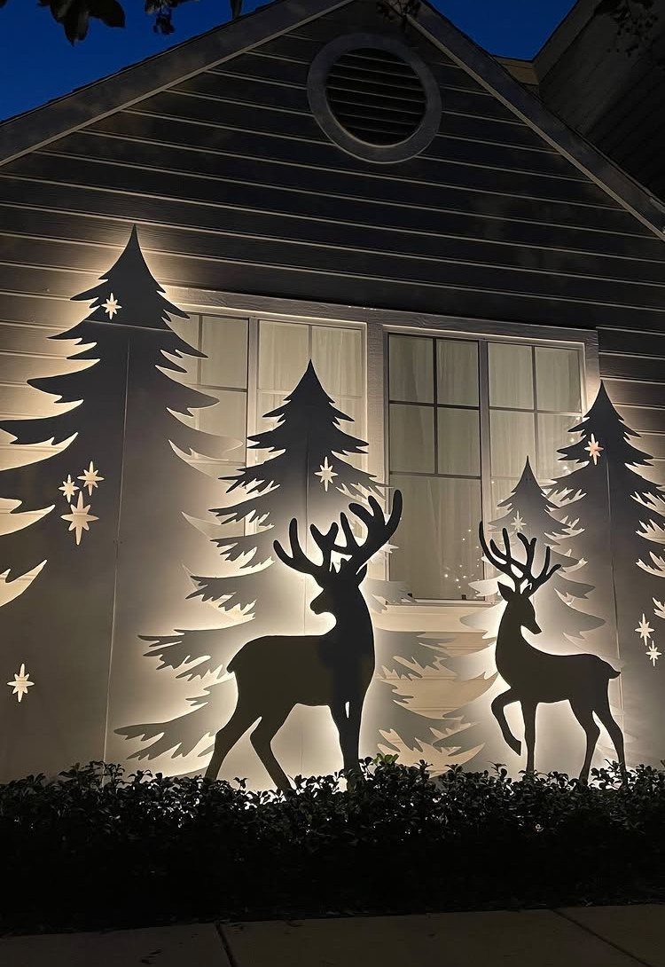 White silhouette cutouts of reindeer and pine trees glowing in front of a window at night.