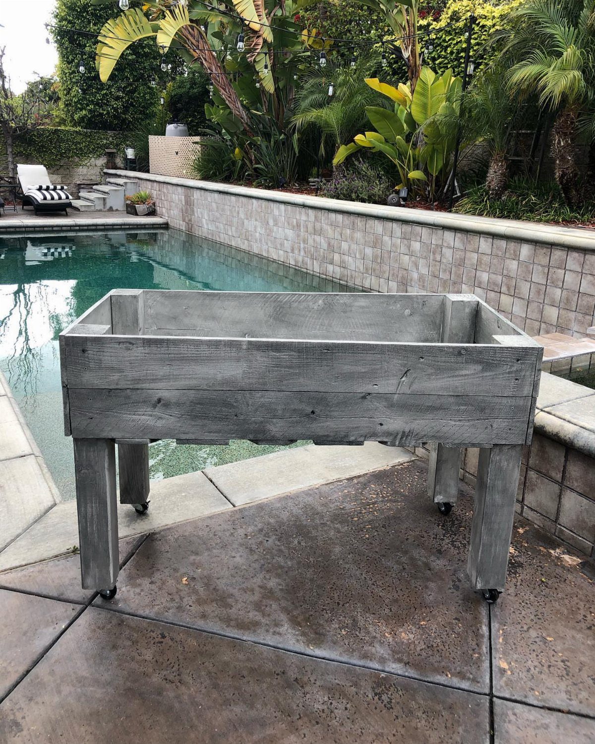 9 Easy DIY Planter Ideas for Wood, Upcycled & Clay Projects 1 Grey wooden planter box with wheels sitting on a patio next to a swimming pool.