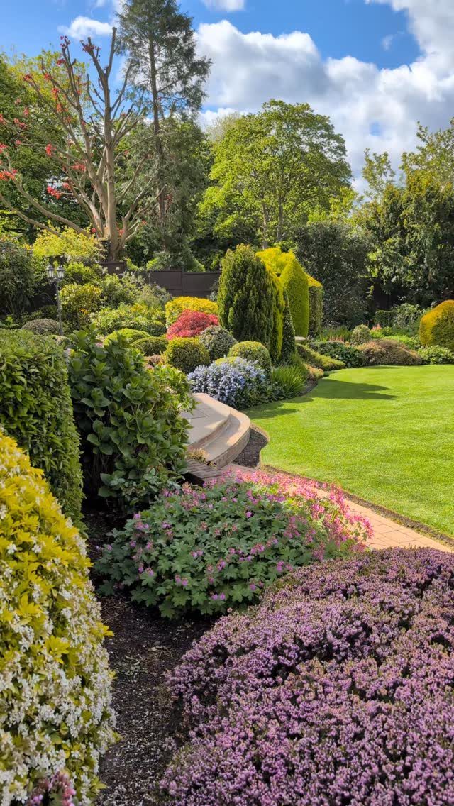 15 Huge Garden Design Ideas for Birds, Light, Night Paths and Still Water 12 A large, landscaped garden with colorful flowering shrubs, green lawns, and tall trees under a blue sky.