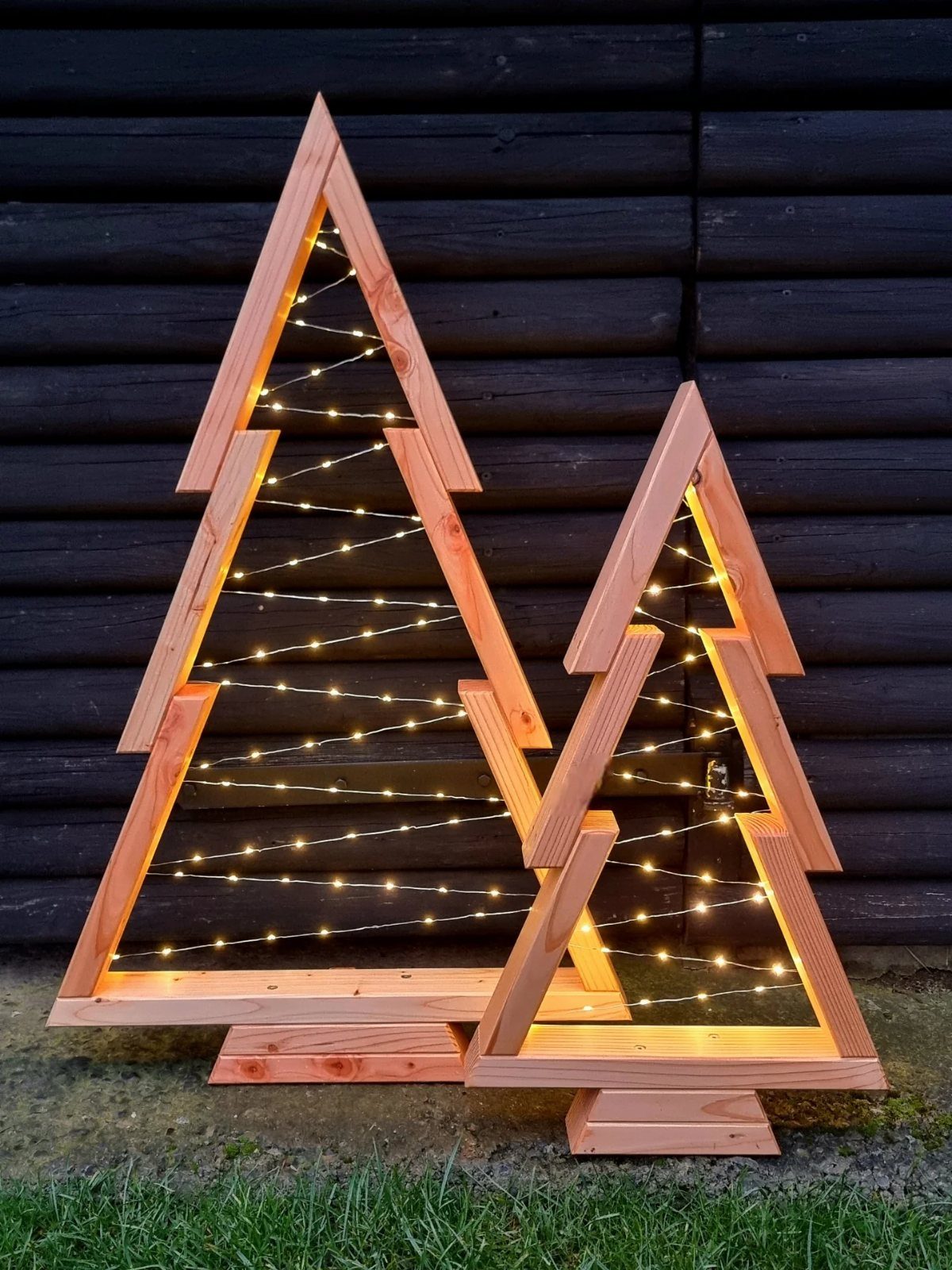 Make Wooden Christmas Trees: 7 DIY Projects from Scrap to Yard 3 Two wooden Christmas tree frames with warm fairy lights standing against a dark wooden wall.