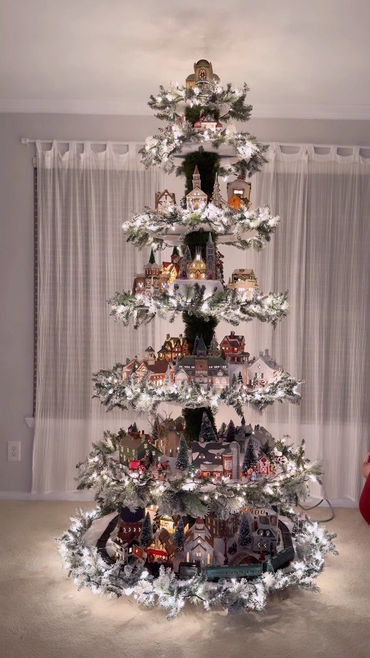 21 Most Beautiful Christmas Trees for Bold and Cozy Looks 5 A snowy Christmas tree decorated with miniature glowing Christmas village houses on multiple tiers.