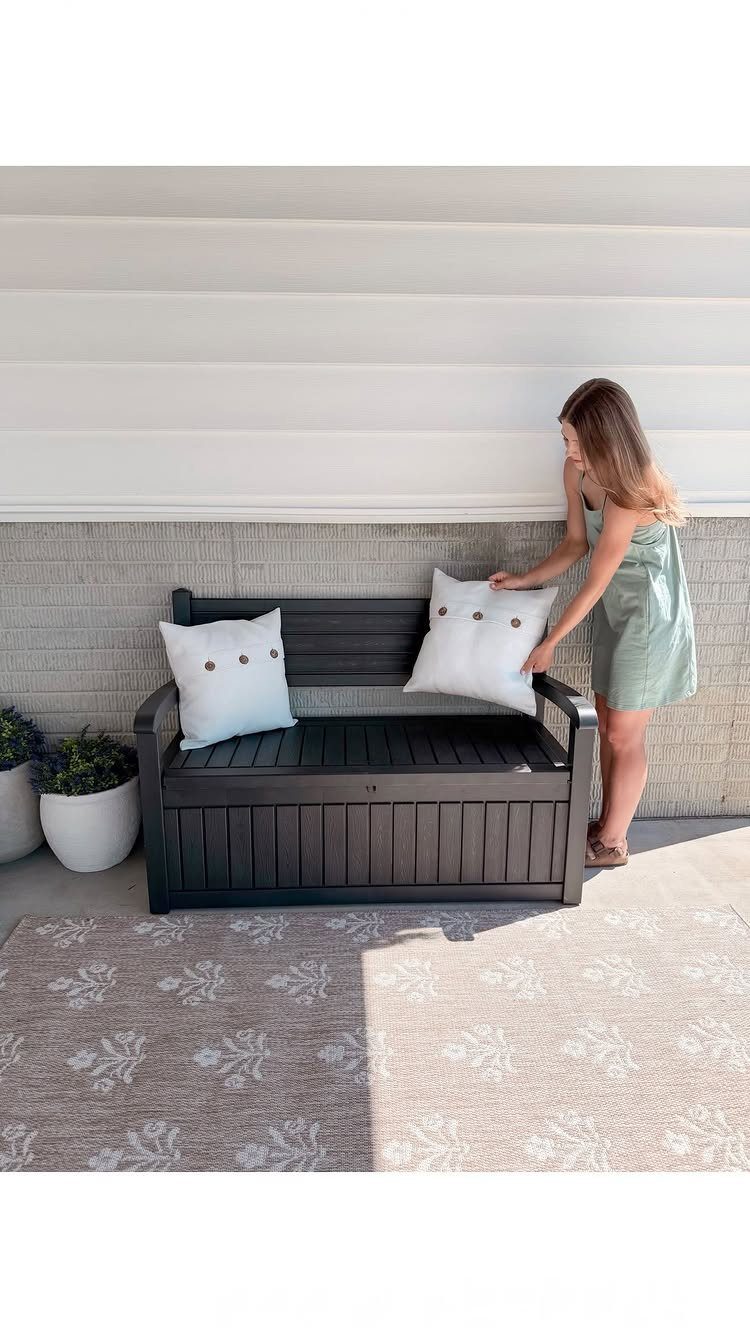 12 Small Backyard Design Ideas for Outdoor Living 4 A woman arranging white pillows on a dark grey wooden outdoor storage bench.