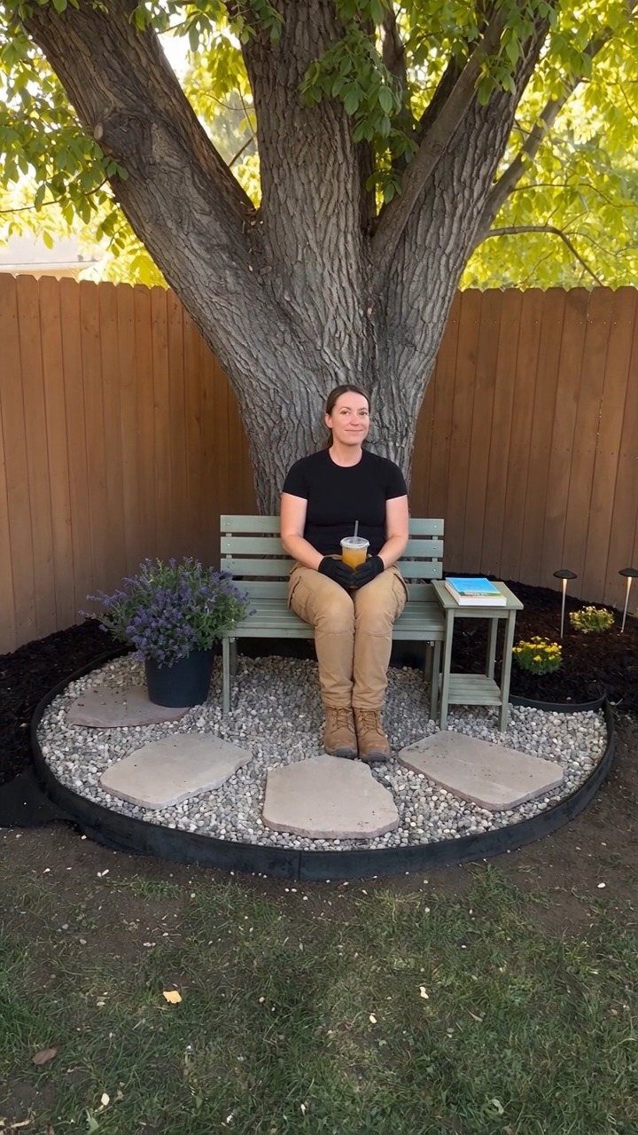 12 Small Backyard Design Ideas for Outdoor Living 6 A woman sitting on a small green wooden bench under a large tree in a garden.