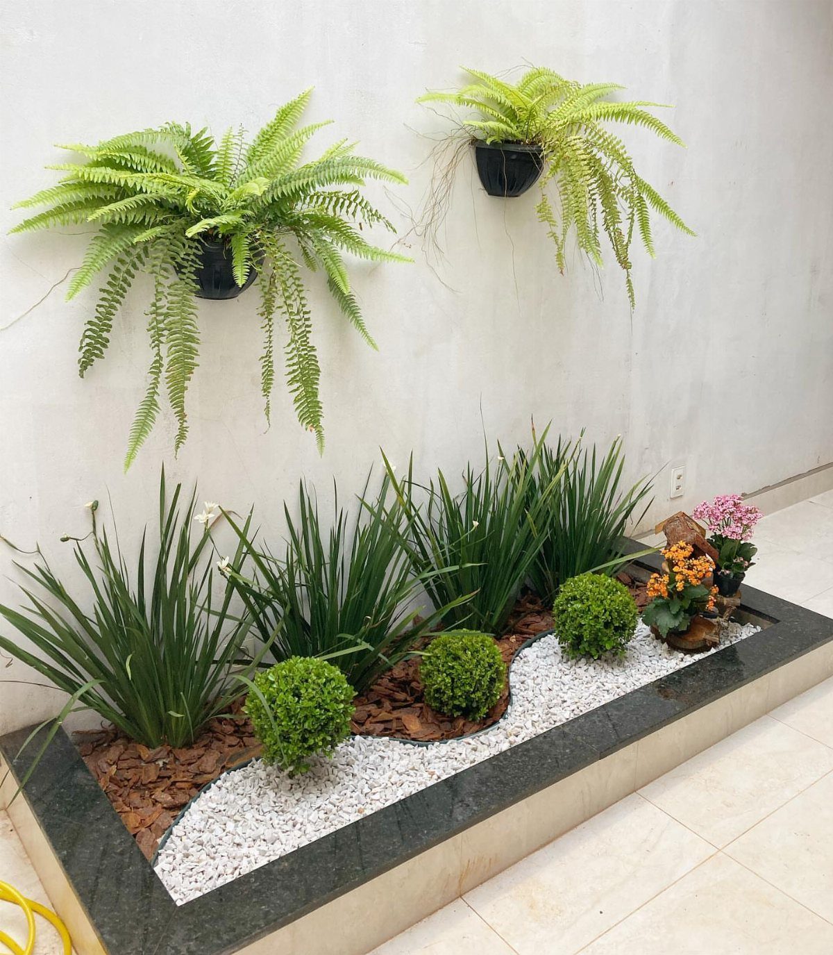 12 Small Backyard Design Ideas for Outdoor Living 2 Small green shrubs and tall grass-like plants arranged in a narrow garden bed with white pebbles.