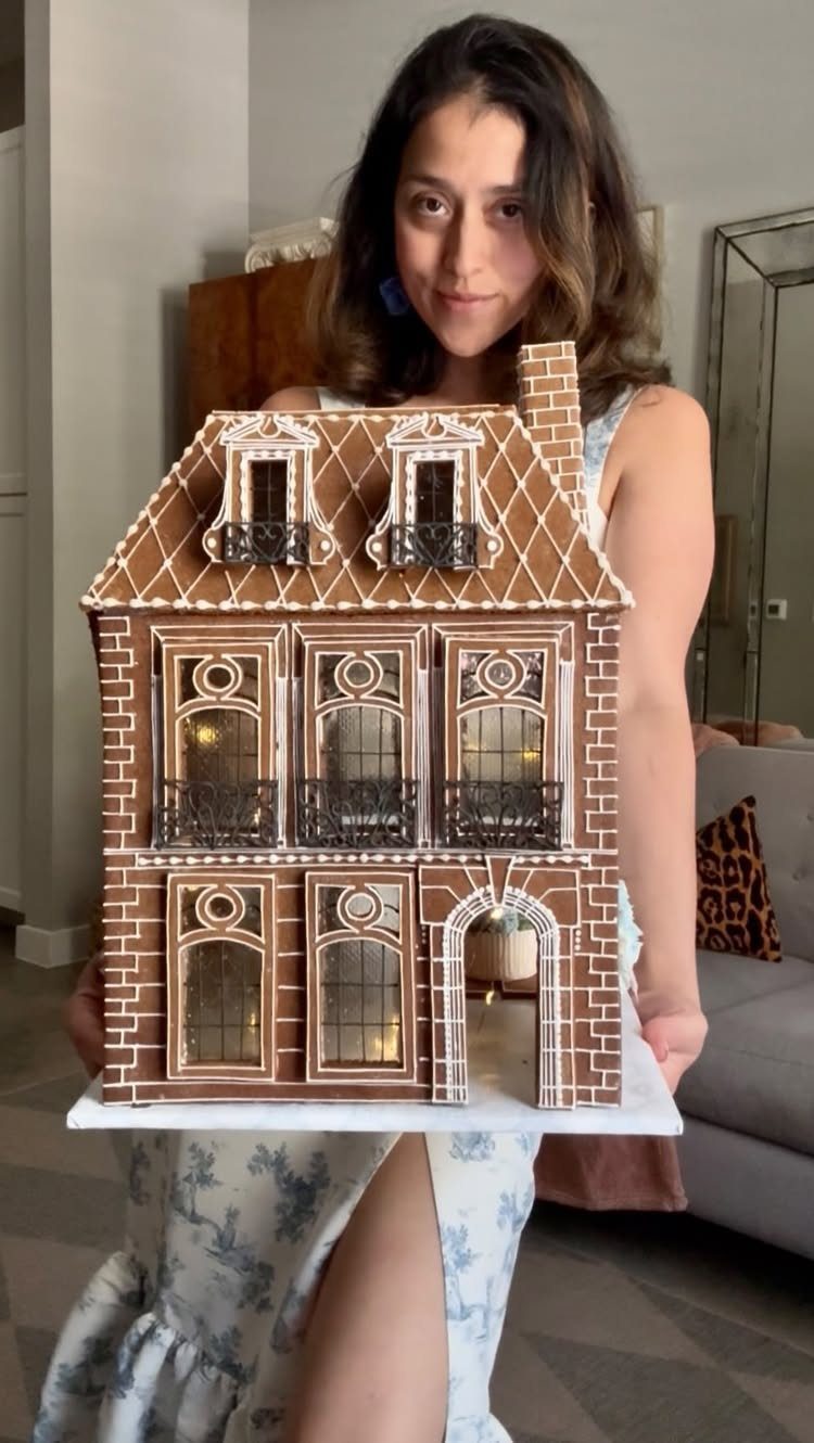 21 Things to Do for Christmas 2026 2 A woman smiling while holding a large, intricately decorated gingerbread house with white icing details.